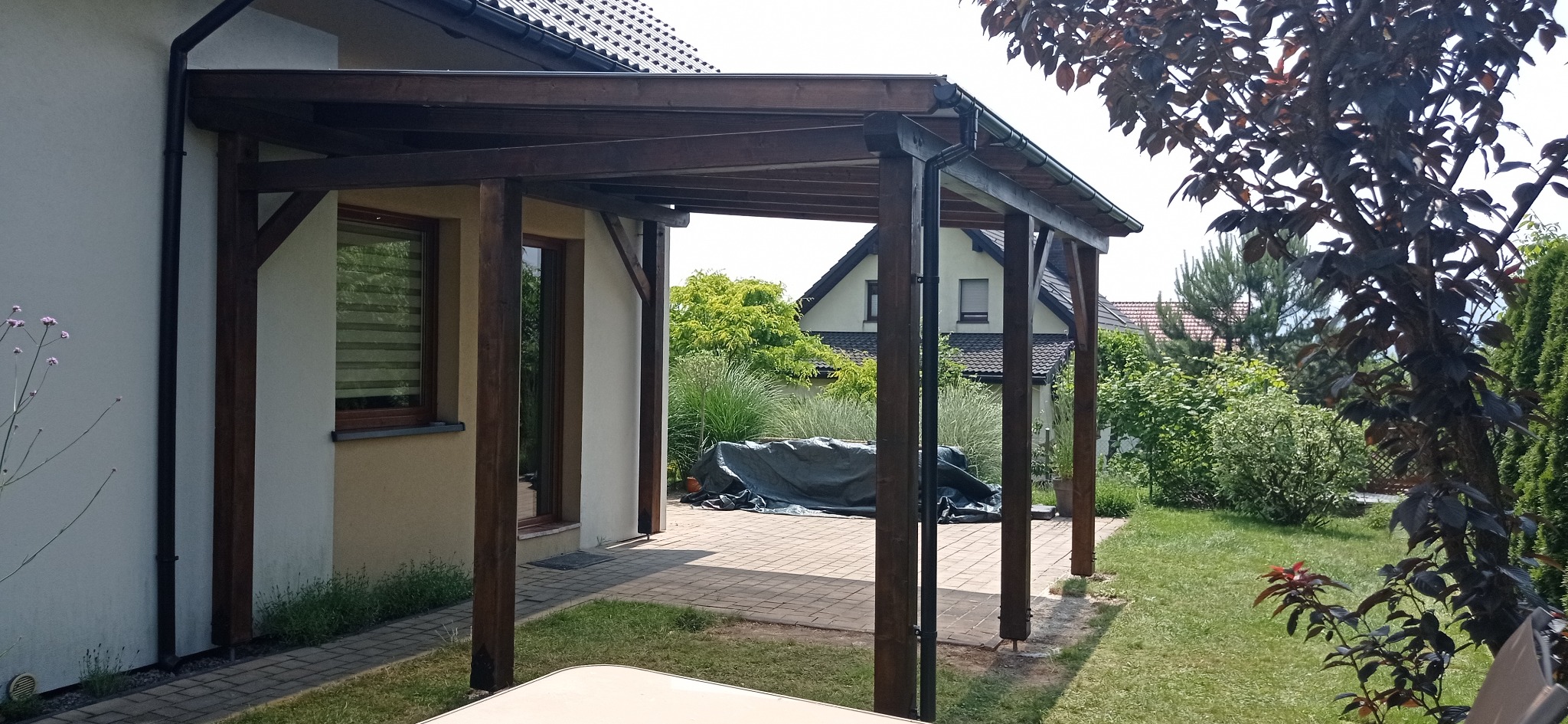 Drewniana konstrukcja z ciemnego drewna, prawdopodobnie zadaszenie tarasu lub pergola, przylegająca do budynku z jasną elewacją, widok na ogród z zielenią i dom w tle.
