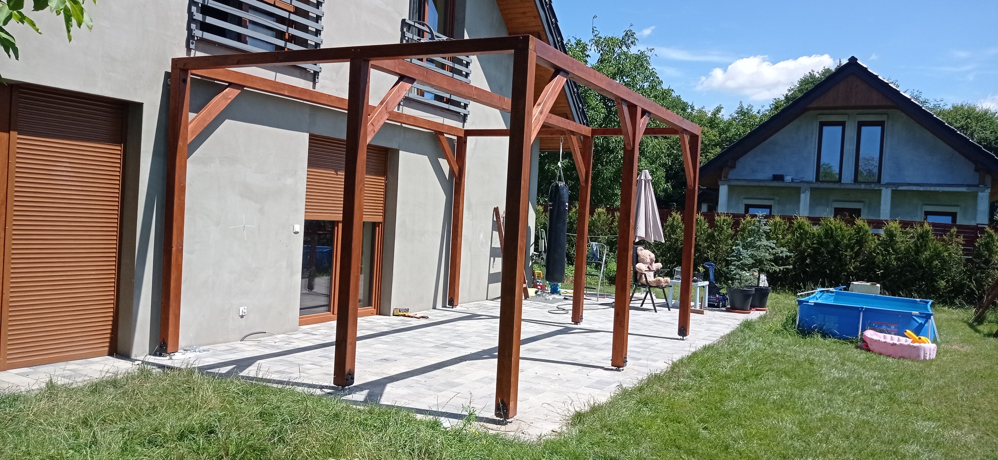 Surowa drewniana pergola w trakcie budowy na wybrukowanym tarasie, z widocznym workiem treningowym, parasolem i basenem dziecięcym w tle.