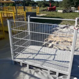 Metalowa klatka pomalowana na szaro, z roboczymi rękawicami zawieszonymi na górnej krawędzi, otoczona żółtymi konstrukcjami i sprzętem budowlanym na zewnątrz.