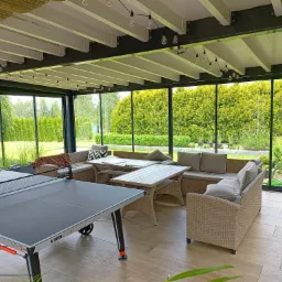 Zadaszony taras z meblami wypoczynkowymi z rattanu, stołem i stołem do ping-ponga; widok na zielony ogród przez przeszklone ściany.