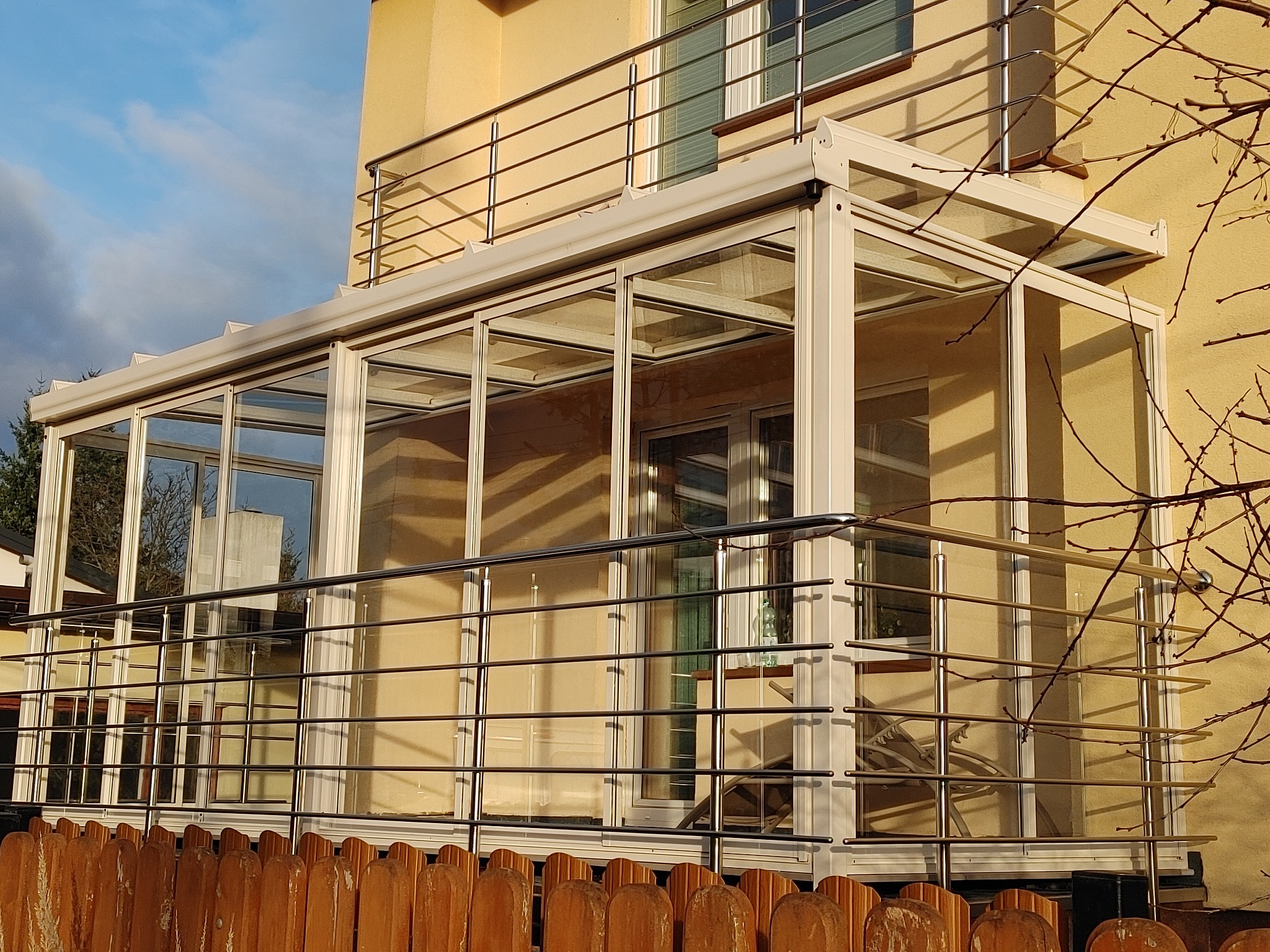 Szklana zabudowa balkonu z białymi ramami i metalową balustradą, widok z zewnątrz, z brązowym drewnianym płotem w pierwszym planie i fragmentem ściany budynku w tle.