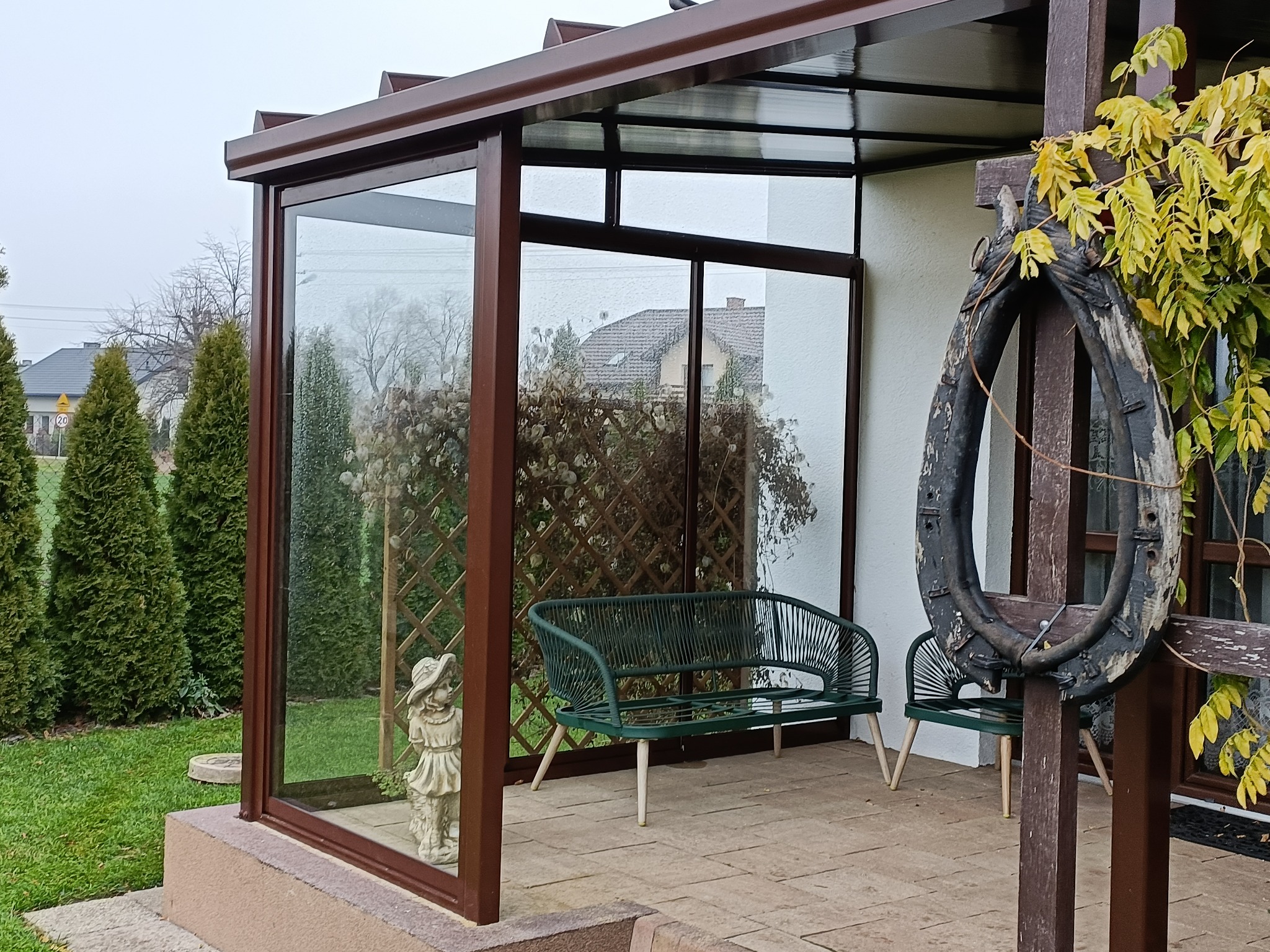 Zadaszony taras z brązową ramą, przeszklonymi ścianami, zieloną ławką i dekoracyjnym, starym chomątem na tle trawnika i zabudowań.