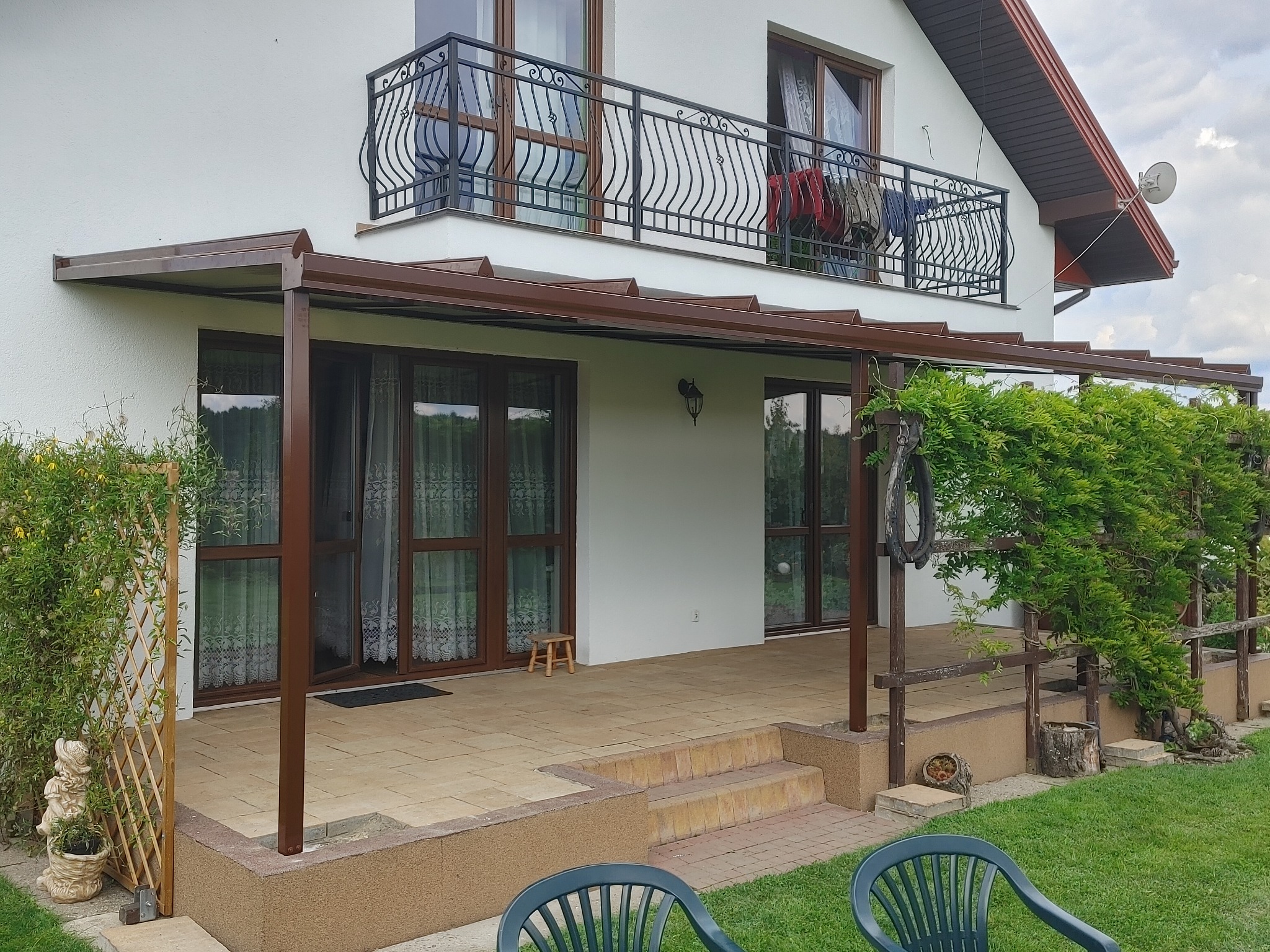 Brązowa pergola metalowa przylegająca do białego domu z balkonem, z roślinami pnącymi po bokach i wyłożonym płytkami tarasem.