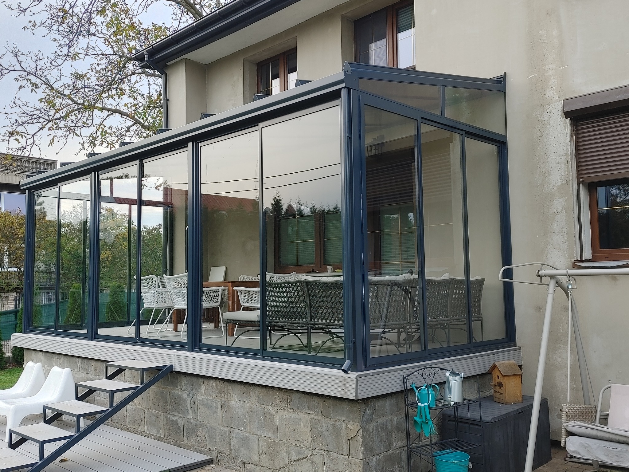 Szklana konstrukcja dobudowana do domu z widocznymi meblami ogrodowymi wewnątrz, schodami wejściowymi i elementami dekoracyjnymi na zewnątrz.