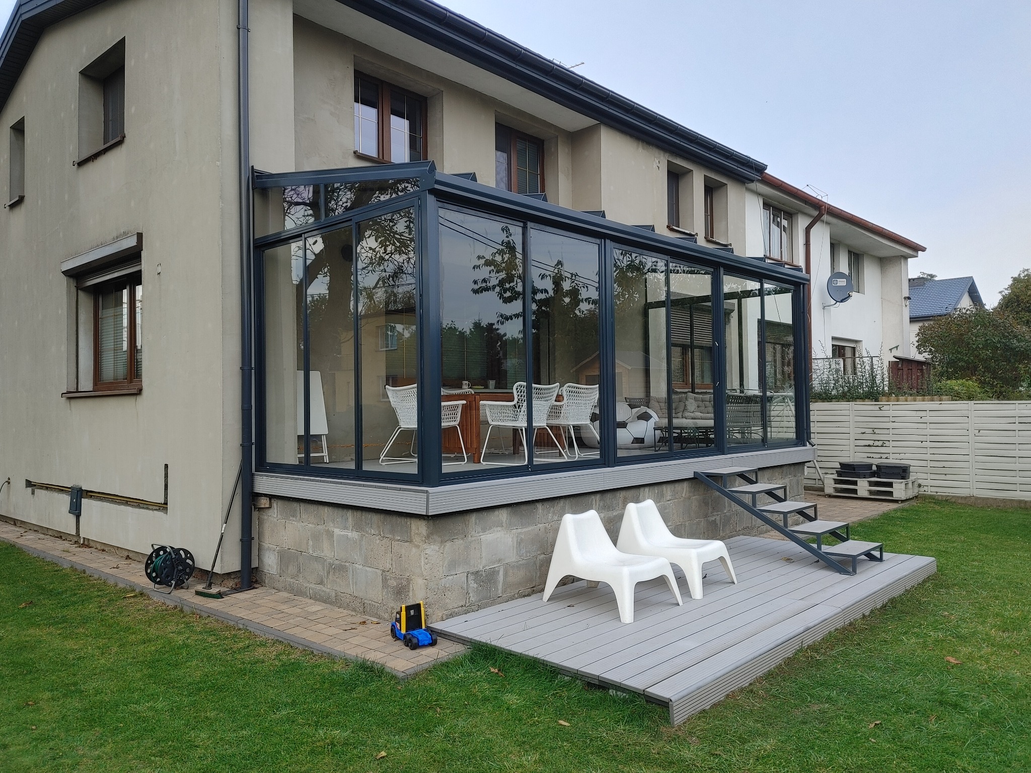 Szklana dobudówka do domu z tarasem z desek kompozytowych, schodami i białymi krzesłami. Na trawniku stoi zabawkowy wózek widłowy.