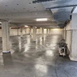 Świeżo wylana, mokra posadzka przemysłowa w hali z betonowymi kolumnami i instalacjami pod sufitem, zacierana za pomocą maszyny do zacierania betonu.