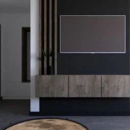 Nowoczesny salon z telewizorem wbudowanym w ścianę z drewnianymi panelami, minimalistyczna komoda pod telewizorem i dekoracyjna roślina w czarnej doniczce, widoczny fragment biurka i krzesła.