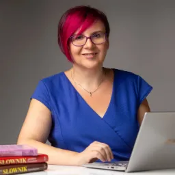 Kobieta z różowymi włosami w niebieskiej sukience pracuje na laptopie, obok stos słowników.