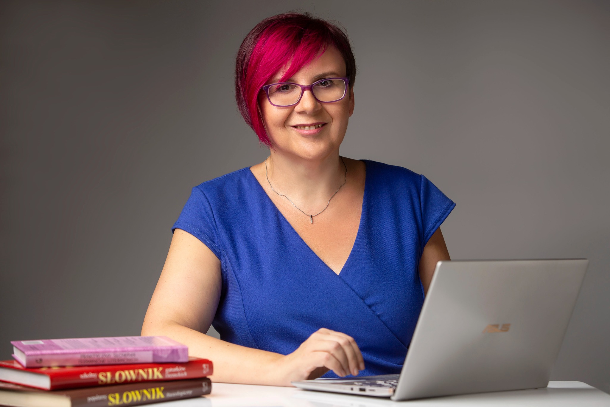 Kobieta z różowymi włosami w niebieskiej sukience pracuje na laptopie, obok stos słowników.