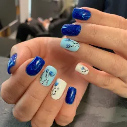 Dłonie z manicure hybrydowym: błyszczące, granatowe paznokcie oraz jasnoniebieskie i białe z ręcznie malowanymi kwiatami.