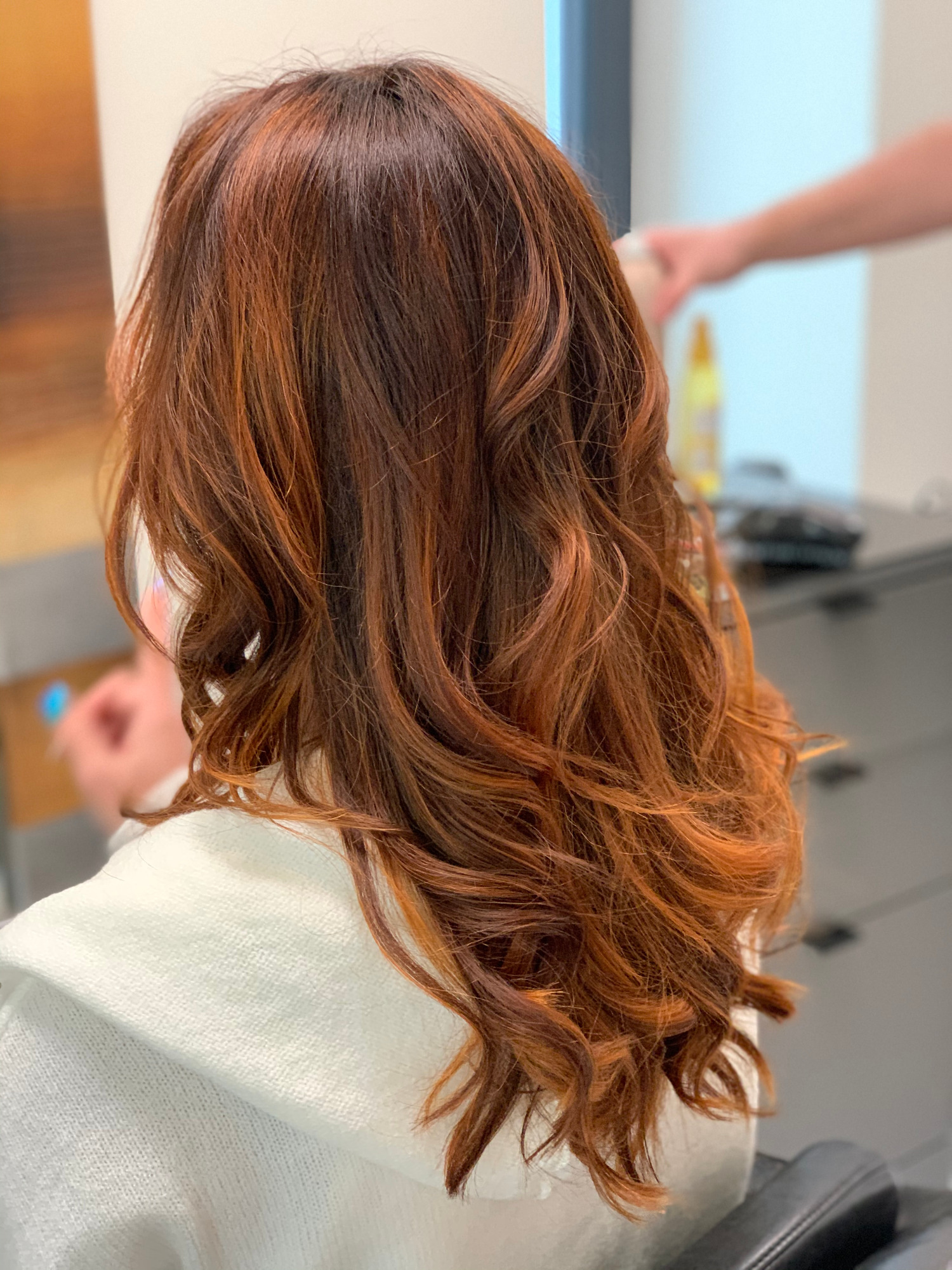 Miedziane, falowane włosy po koloryzacji, widok z tyłu, na karku biały peleryna fryzjerska, w tle rozmyte stanowisko pracy.