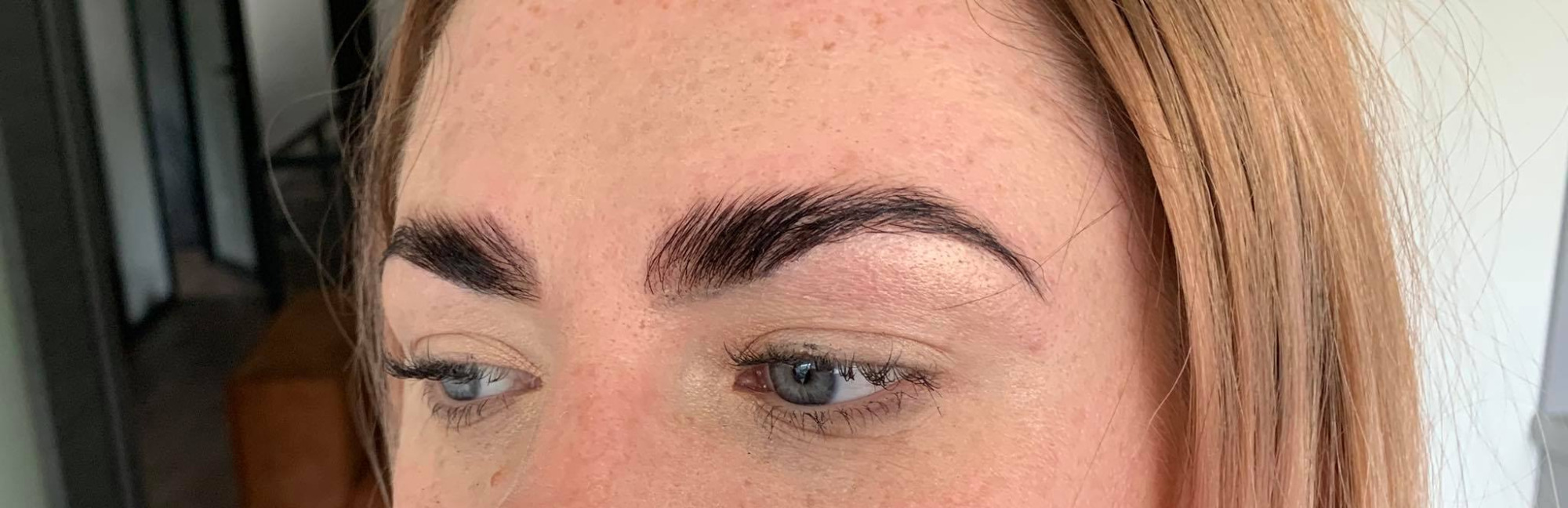 Zbliżenie na kobiece oko z wyraźnie podkreślonymi, ciemnymi brwiami po zabiegu laminacji, widoczne drobne piegi na skórze i jasne włosy.