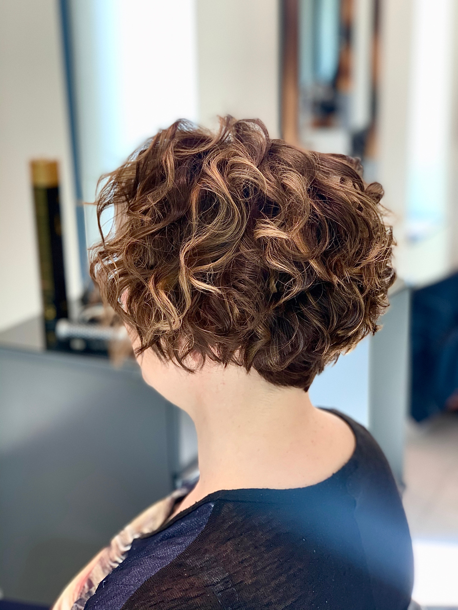 Krótkie, brązowe, kręcone włosy z blond pasemkami, widok z tyłu, w tle fragment salonu.