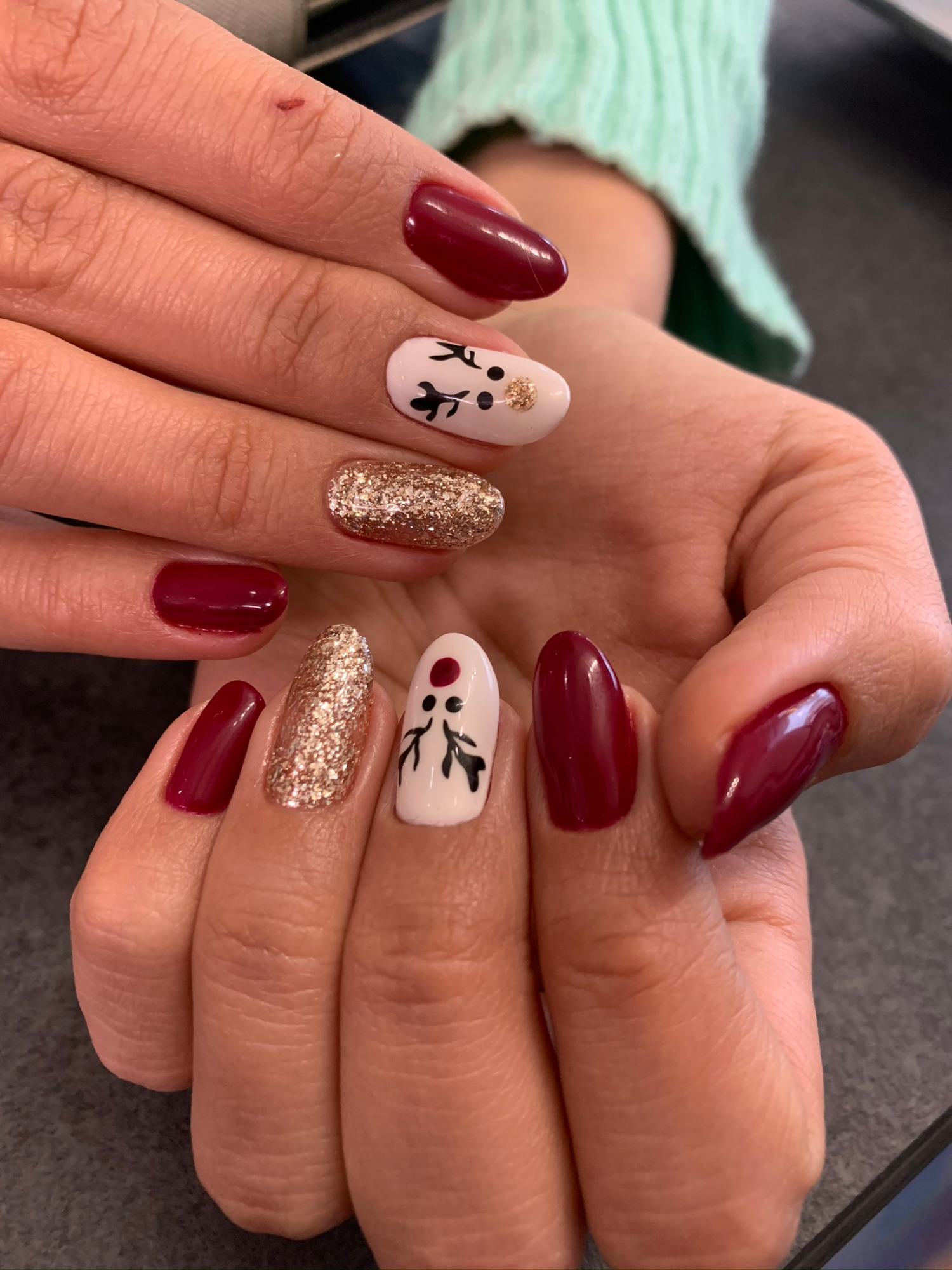 Dłonie z manicure hybrydowym: paznokcie w kolorze burgundu, złoty brokat i białe z czarnym wzorem gałązek i złotymi kropkami.