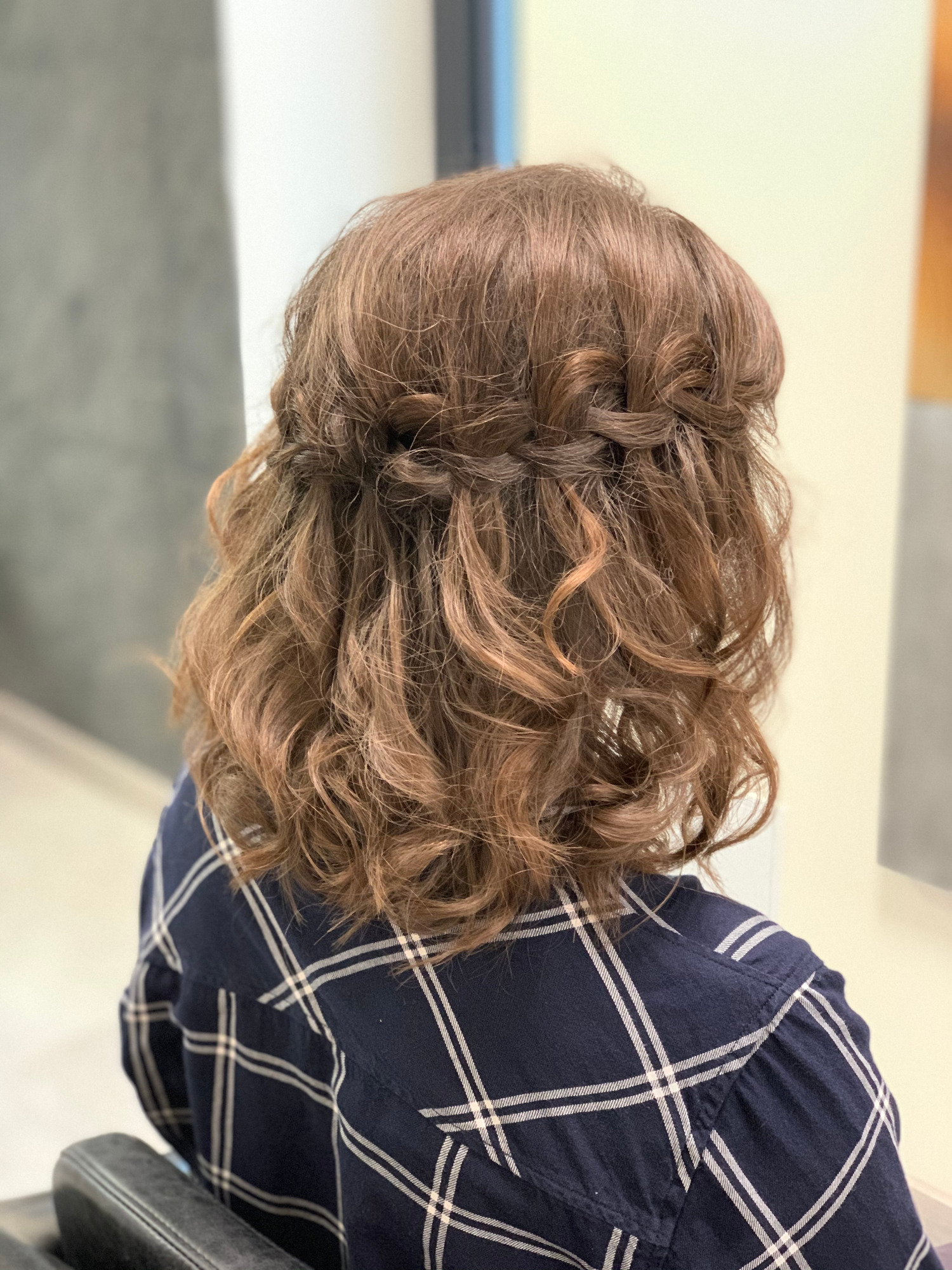 Kobieta z brązowymi włosami upiętymi w warkocz i luźne loki, widok z tyłu, ubrana w koszulę w kratę.