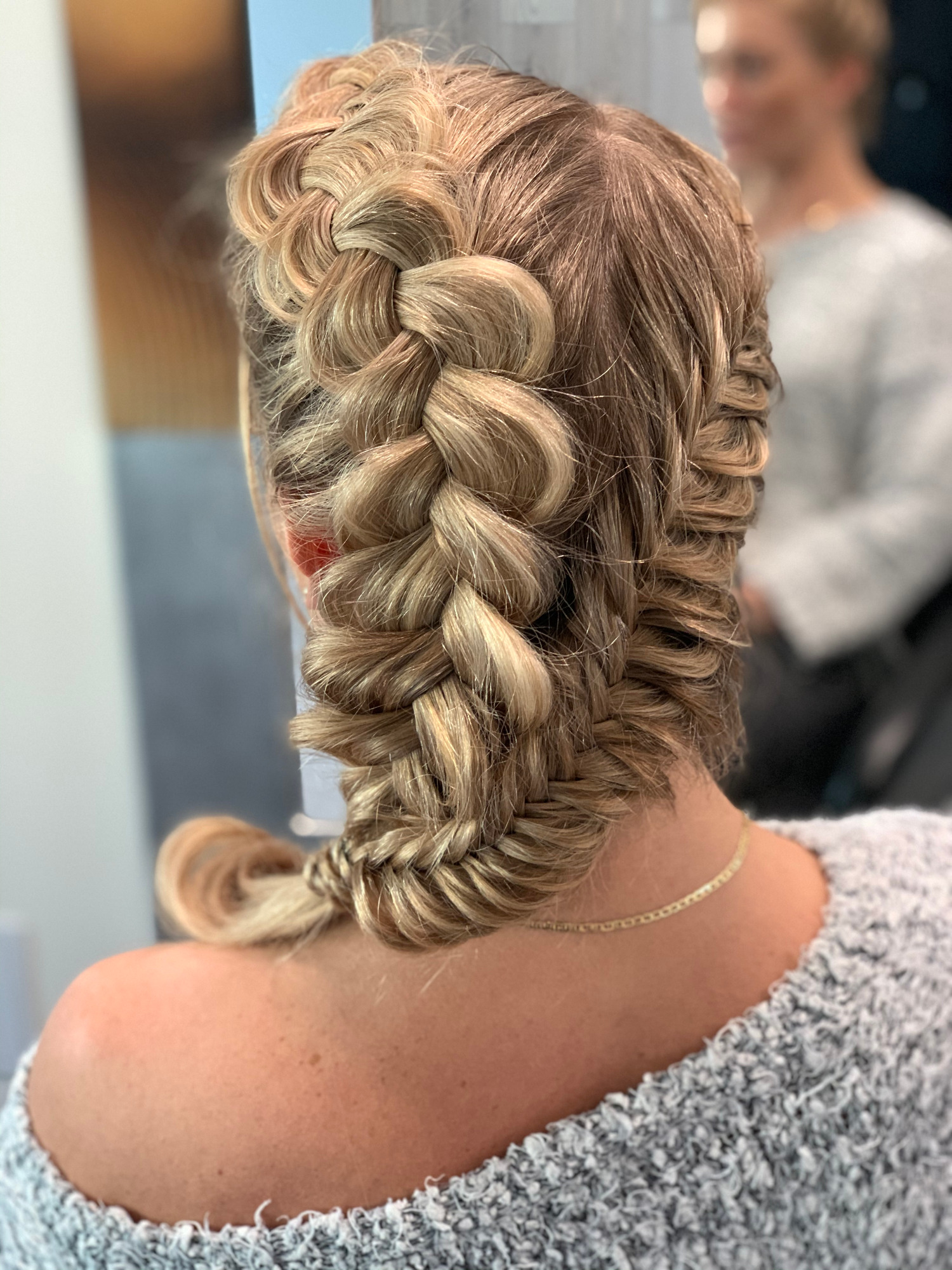 Tył głowy kobiety z zaplecionym, blond warkoczem dobieranym, widoczny złoty łańcuszek na szyi i fragment szarego swetra.