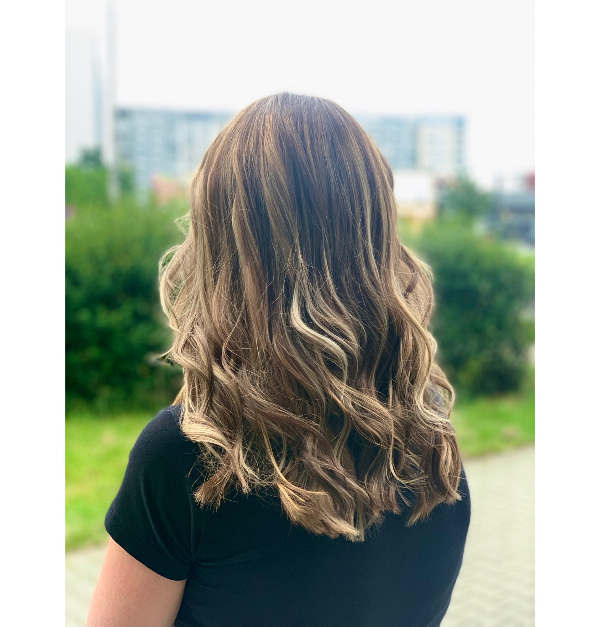 Kobieta z tyłu, prezentująca brązowe włosy z blond pasemkami, delikatnie pofalowane i ułożone w luźne loki, na rozmytym tle zieleni i miejskiego krajobrazu.