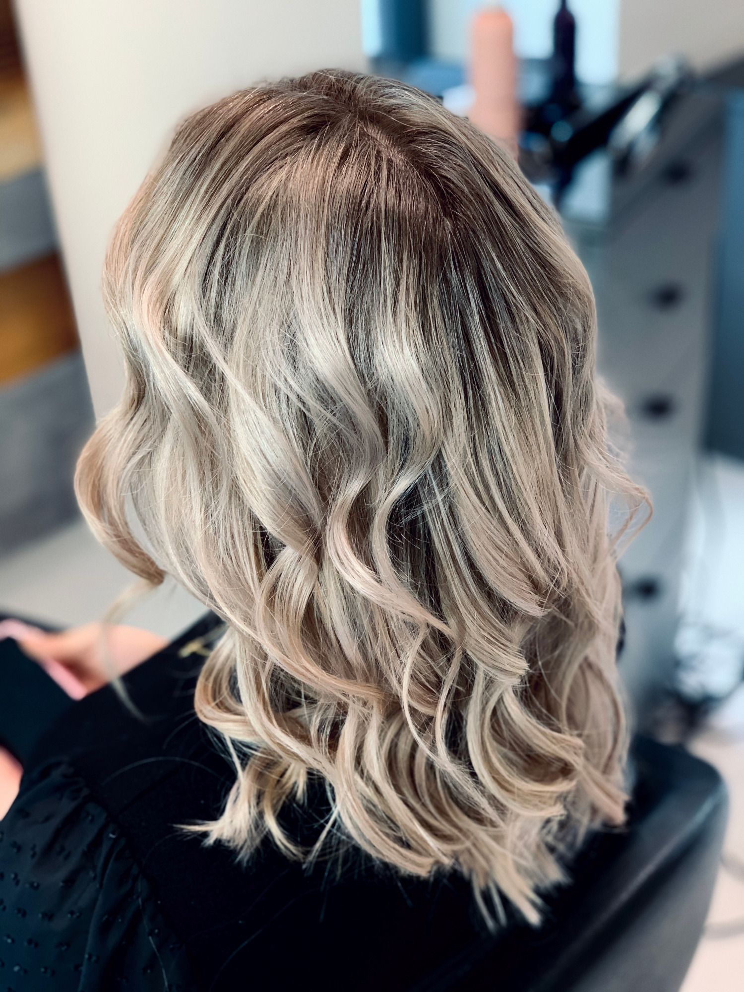 Tył głowy kobiety z blond włosami w odcieniach beżu i platyny, delikatnie pofalowanymi, w salonie fryzjerskim.