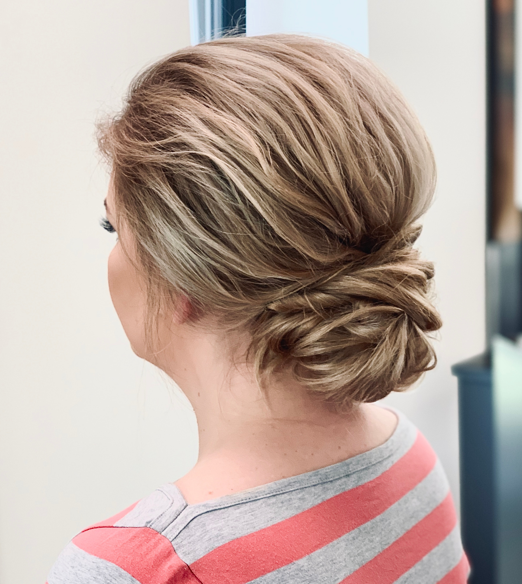 Upięcie włosów blond, kok niski z tyłu głowy, widok z boku na kark i fragment ramienia w bluzce w szaro-różowe paski.