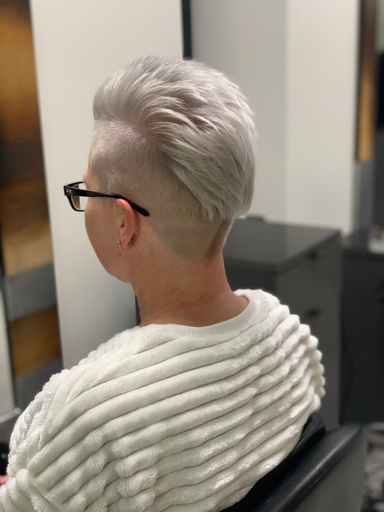 Krótkie, platynowe włosy z podgoleniem na karku, widok z tyłu, kobieta w okularach i białym swetrze.