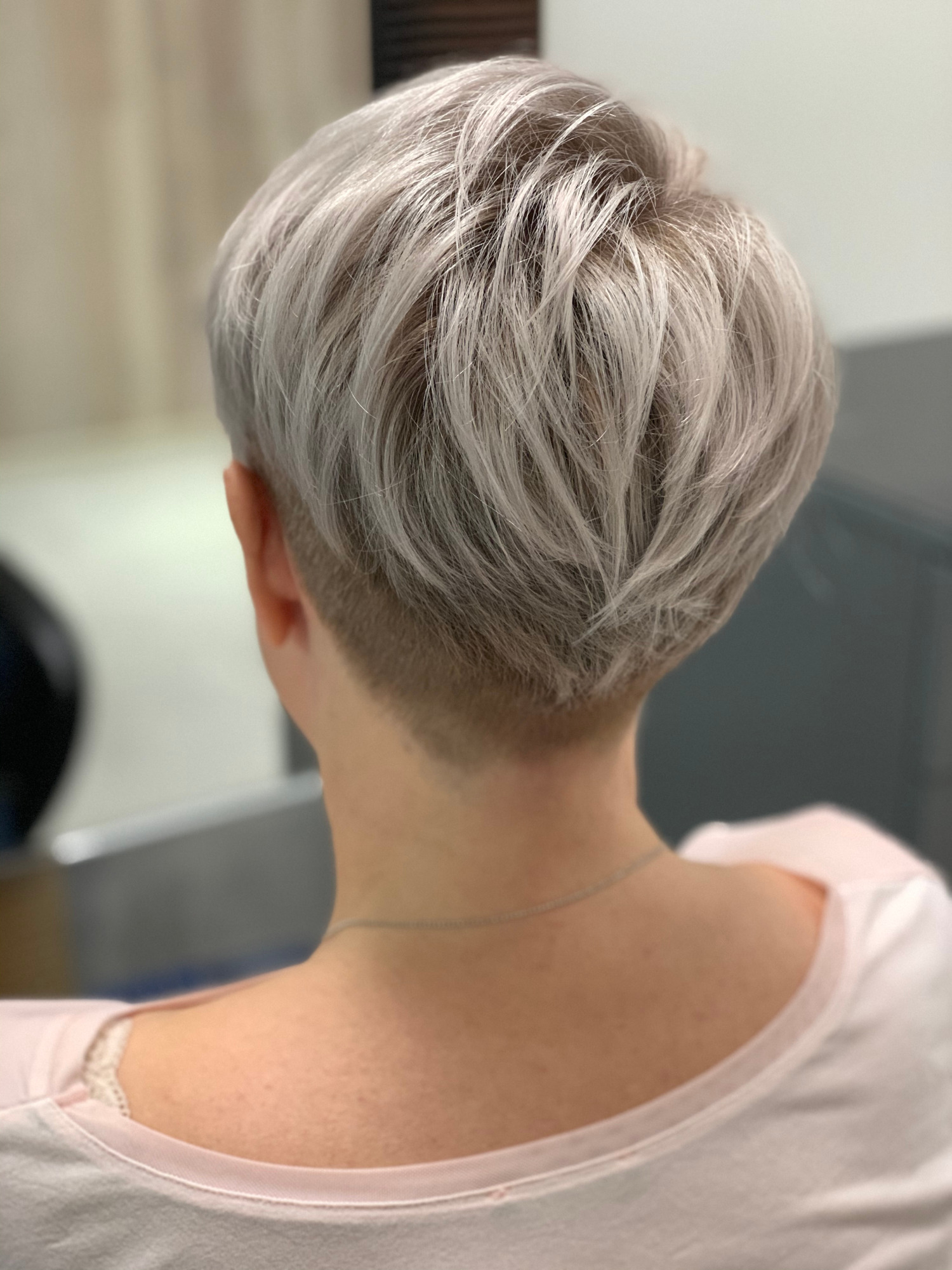 Krótkie, platynowe włosy z podgoleniem na karku, widok z tyłu, wykonane w salonie fryzjerskim w Łodzi. Delikatny srebrny łańcuszek na szyi.