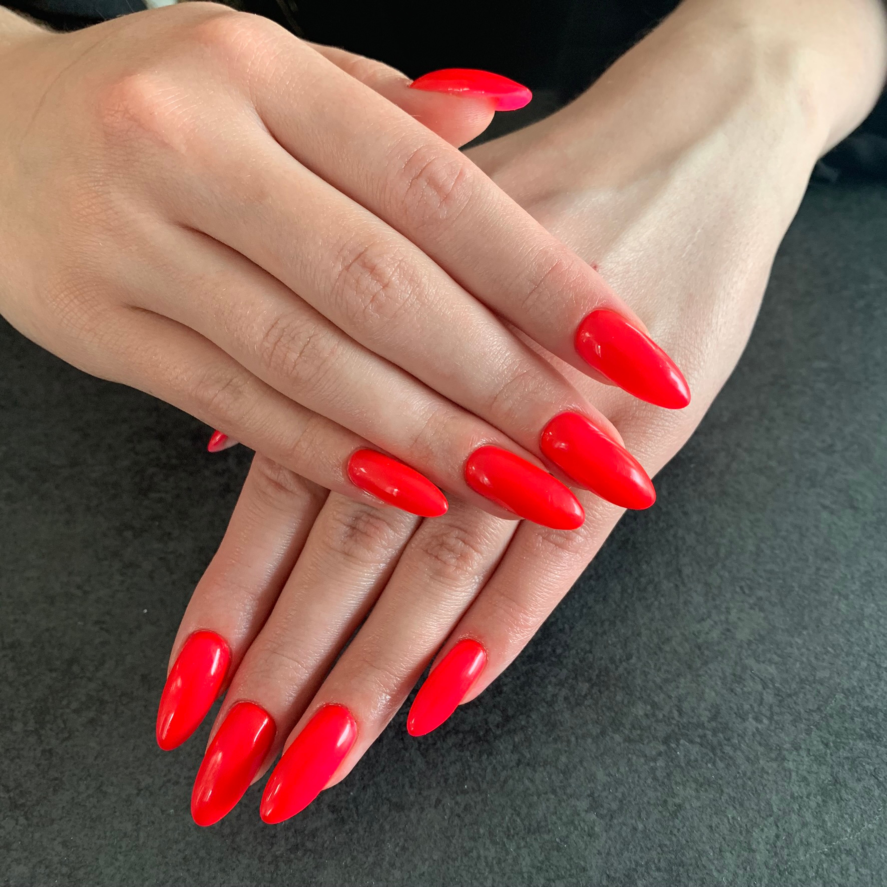 Dłonie z jaskrawoczerwonym manicure hybrydowym, migdałowy kształt paznokci, gładka powierzchnia, na ciemnym tle.