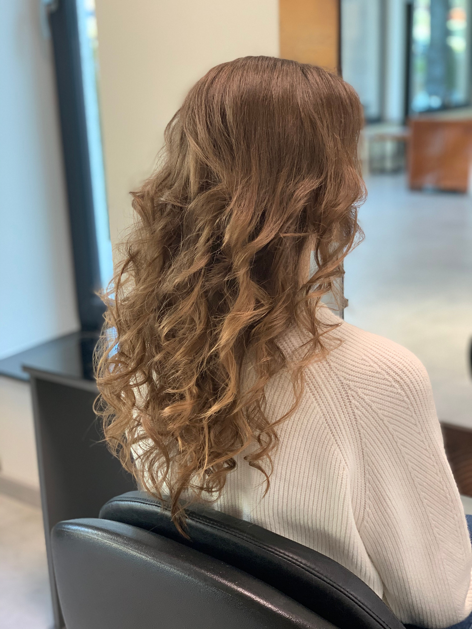 Kobieta siedząca na czarnym krześle, z widocznymi od tyłu brązowymi, kręconymi włosami, w jasnym swetrze, w tle fragment wnętrza salonu.