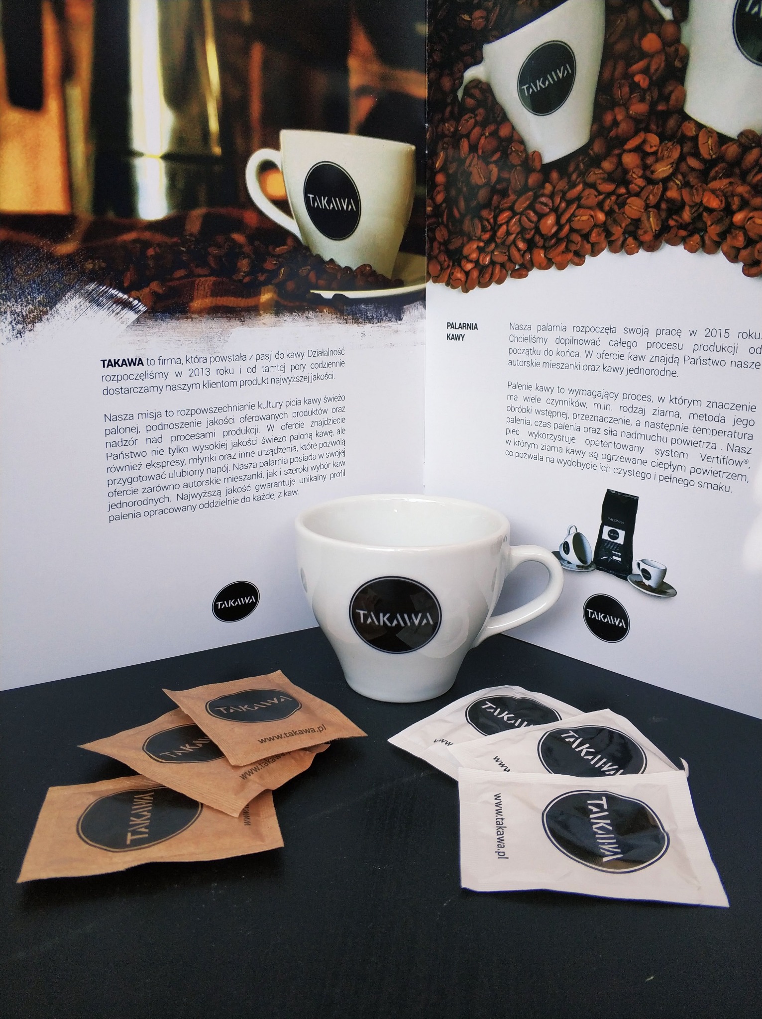 Reklama Palarni Kawy Takawa prezentująca filiżanki z logo, ziarna kawy oraz saszetki z kawą na ciemnym tle.