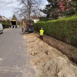 Prace ziemne na ulicy: koparka wykopuje rów, pracownik w żółtej kamizelce nadzoruje, w tle ciężarówka i zabudowa jednorodzinna.