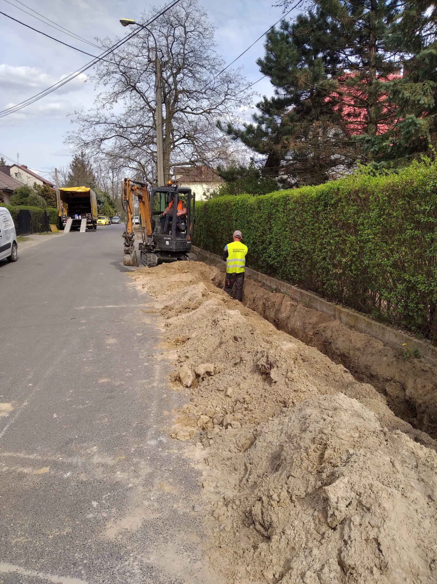 Prace ziemne na ulicy: koparka wykopuje rów, pracownik w żółtej kamizelce nadzoruje, w tle ciężarówka i zabudowa jednorodzinna.
