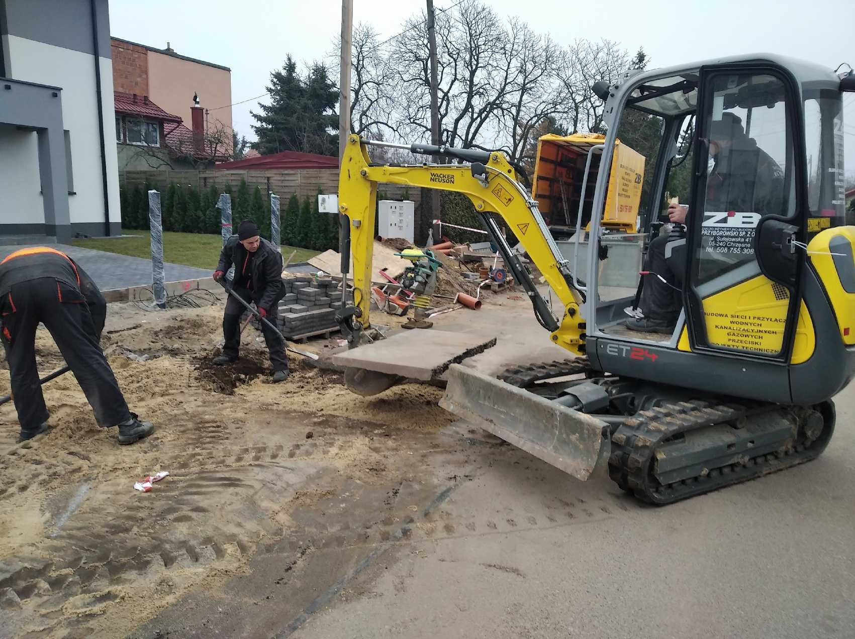 Żółta mini koparka Wacker Neuson ET24 podczas prac ziemnych, dwóch robotników z łopatami przygotowuje teren pod kostkę brukową, widoczny stos kostki i ubijarka w tle.