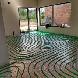 Zielone rury ogrzewania podłogowego ułożone na folii w jasnym pomieszczeniu z dużymi oknami, widok na zewnątrz i fragment ściany z cegły.