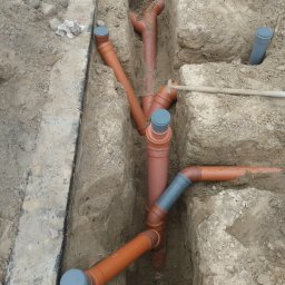 Przedsiębiorstwo Usługowe MAKSTOM - Pomarańczowe rury kanalizacyjne z szarymi złączkami ułożone w wykopie ziemnym, widok z góry. Część rur zakryta ziemią. Na krawędzi wykopu fragment betonowej konstrukcji.