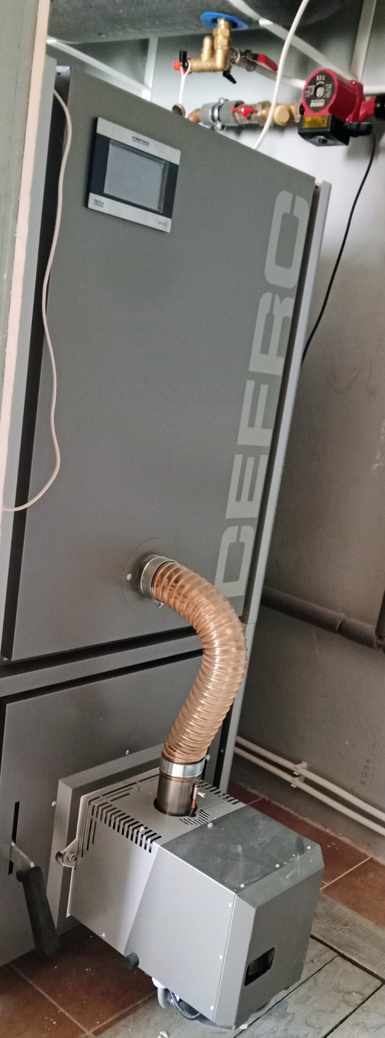 Szary piec CO z panelem sterowania i rurą odprowadzającą spaliny, zainstalowany w kotłowni. Widoczne elementy instalacji na ścianie.