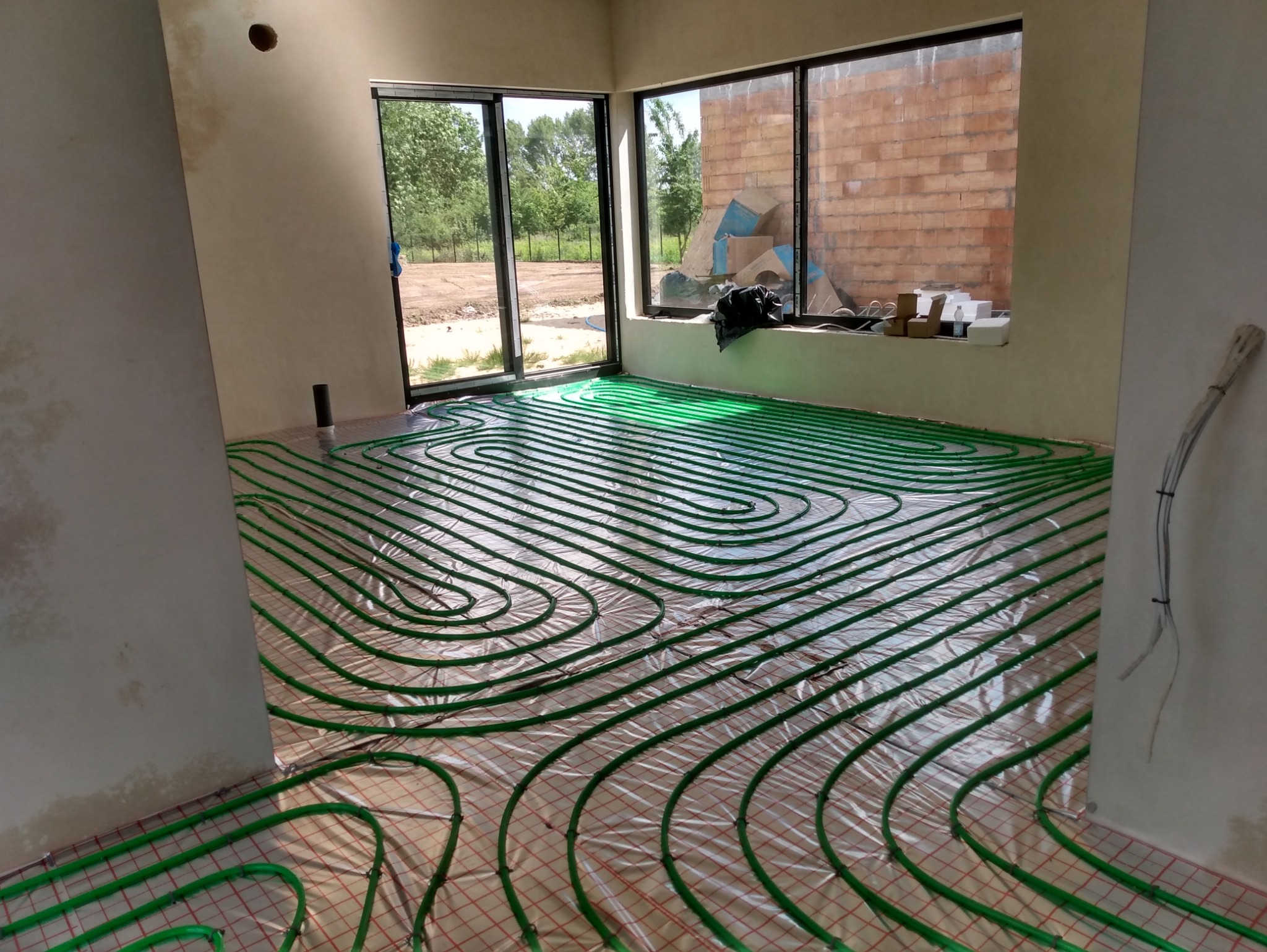 Zielone rury ogrzewania podłogowego ułożone na folii w jasnym pomieszczeniu z dużymi oknami, widok na zewnątrz i fragment ściany z cegły.