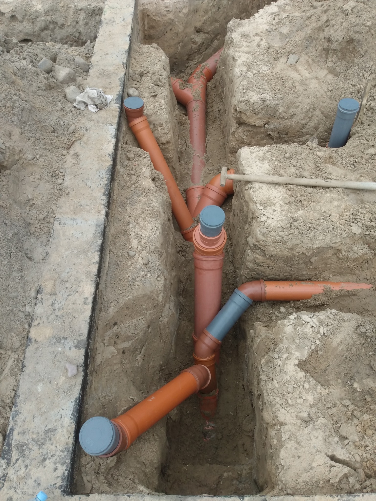 Pomarańczowe rury kanalizacyjne z szarymi złączkami ułożone w wykopie ziemnym, widok z góry. Część rur zakryta ziemią. Na krawędzi wykopu fragment betonowej konstrukcji.