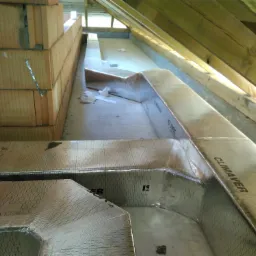 Fragment instalacji wentylacyjnej z izolowanych kanałów, widoczne elementy murowane i konstrukcja dachu, oświetlenie naturalne wpada z góry.