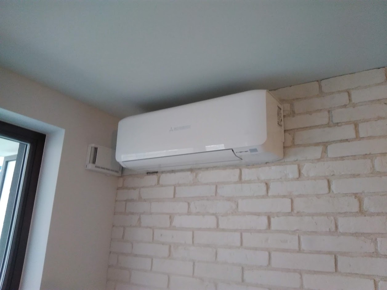 Biały klimatyzator Mitsubishi zamontowany na ścianie z białej cegły w mieszkaniu, widoczny fragment okna.
