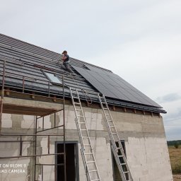 Elektroinstalacje - Instalator w uprzęży montuje panele słoneczne na dachu domu w trakcie budowy, widoczne rusztowanie i drabiny.