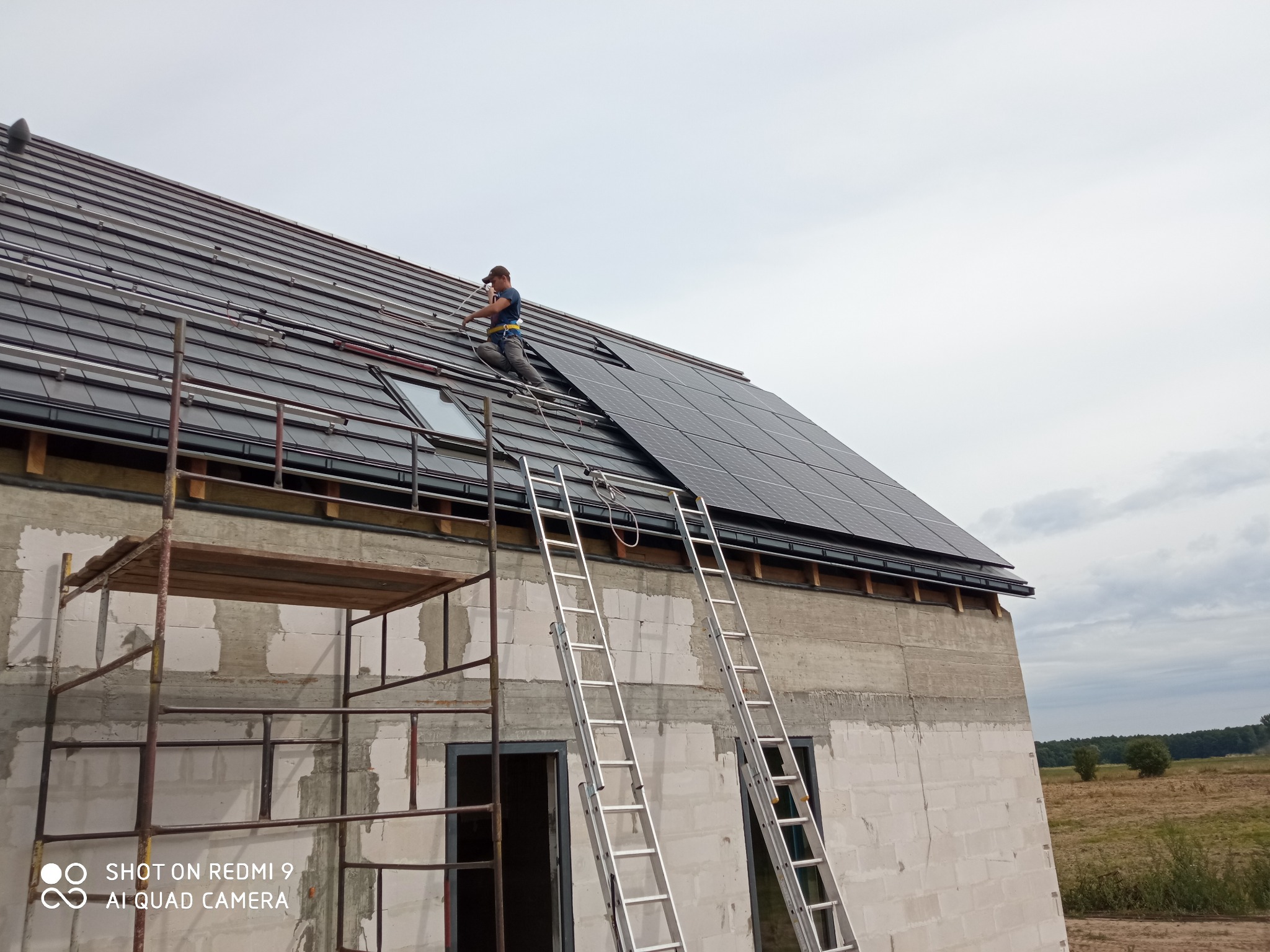 Instalator w uprzęży montuje panele słoneczne na dachu domu w trakcie budowy, widoczne rusztowanie i drabiny.