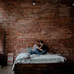 Para siedzi na łóżku na tle ceglanej ściany w loftowym wnętrzu, z widocznym oknem z zasłonami i dekoracyjnym napisem 'Kiss me good night'.