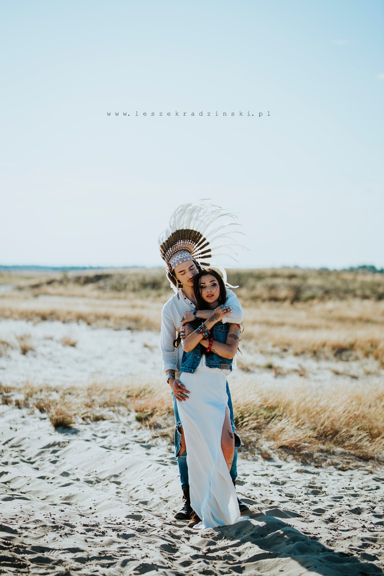 Para w stylizacji indiańskiej na piaszczystym terenie: mężczyzna w pióropuszu obejmuje kobietę w białej sukni z rozcięciem, widoczne tatuaże i biżuteria.