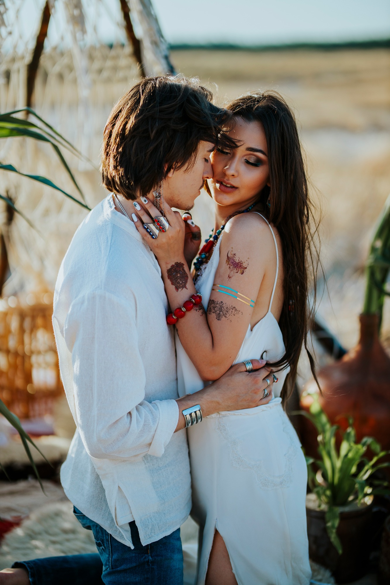 Młoda para w stylu boho, on w białej koszuli i jeansach, ona w białej sukience z rozcięciem, ozdobiona tatuażami i biżuterią, obejmująca się na tle trzcinowej konstrukcji i polnej roślinności.