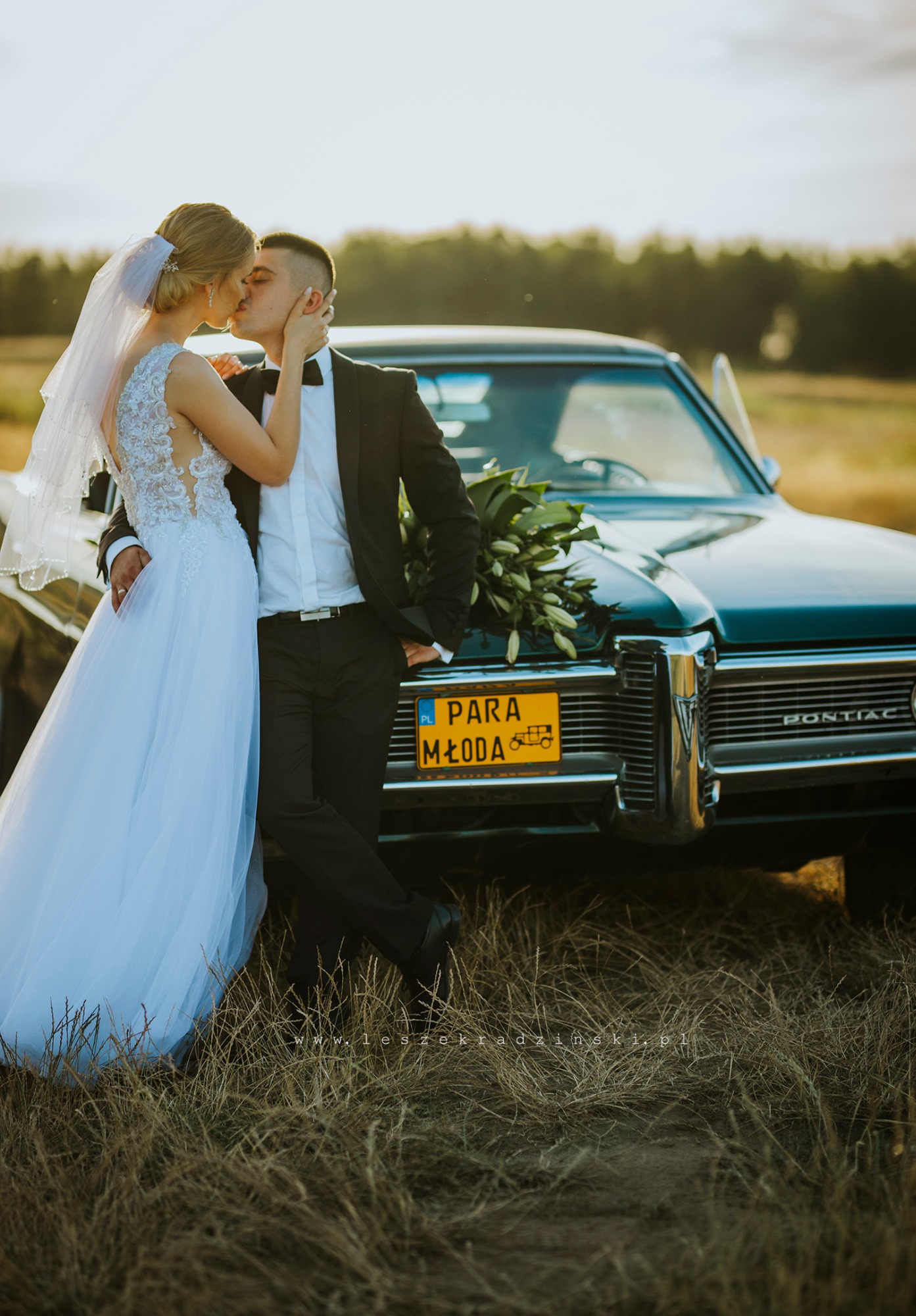 Pocałunek nowożeńców na tle turkusowego Pontiaca z tablicą 'Para Młoda' i bukietem lilii na masce, w plenerze o zachodzie słońca.