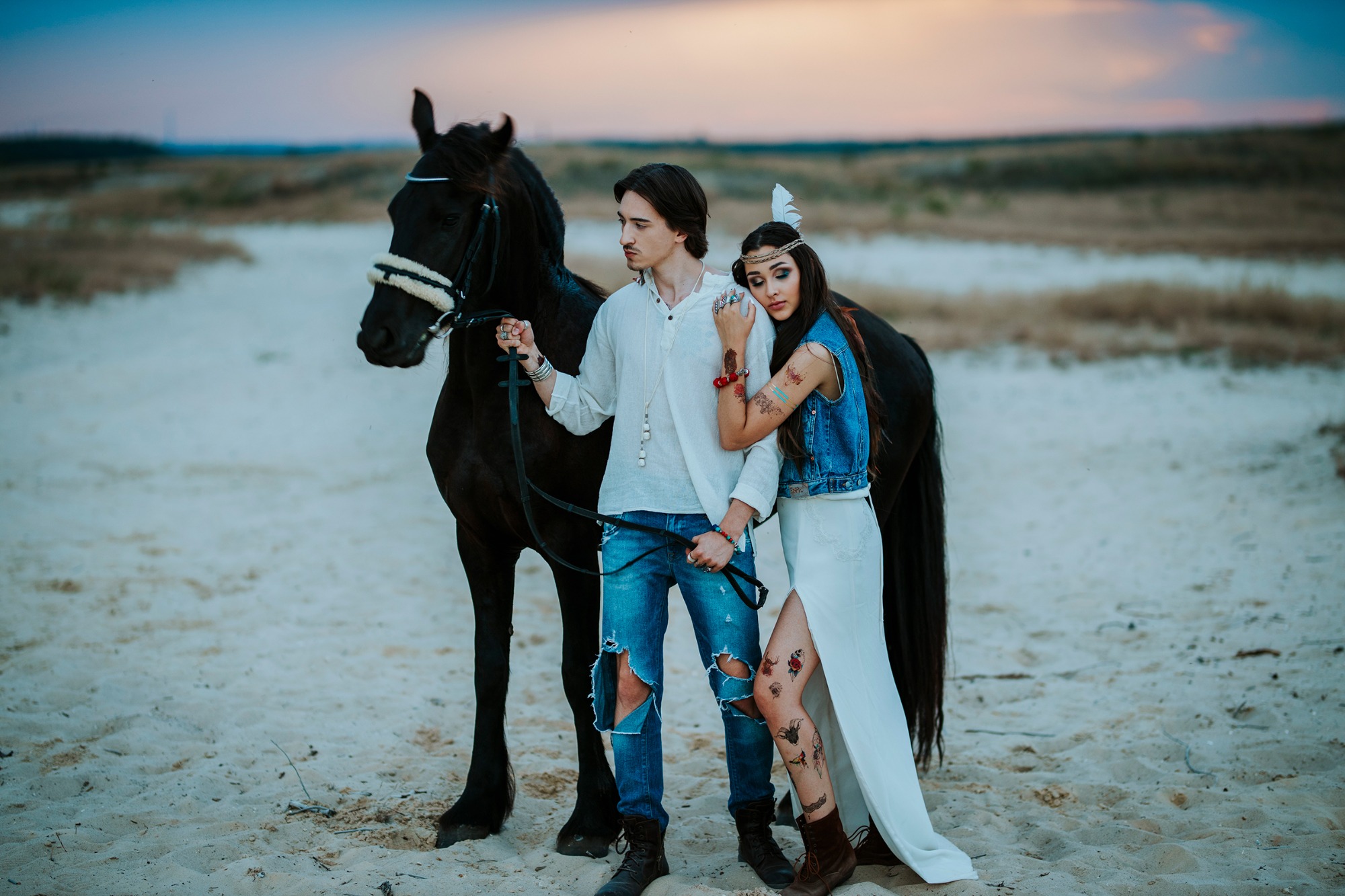 Młoda para w stylu boho z czarnym koniem na piaszczystej plaży o zmierzchu, kobieta z piórem we włosach i tatuażami, mężczyzna w podartych jeansach.