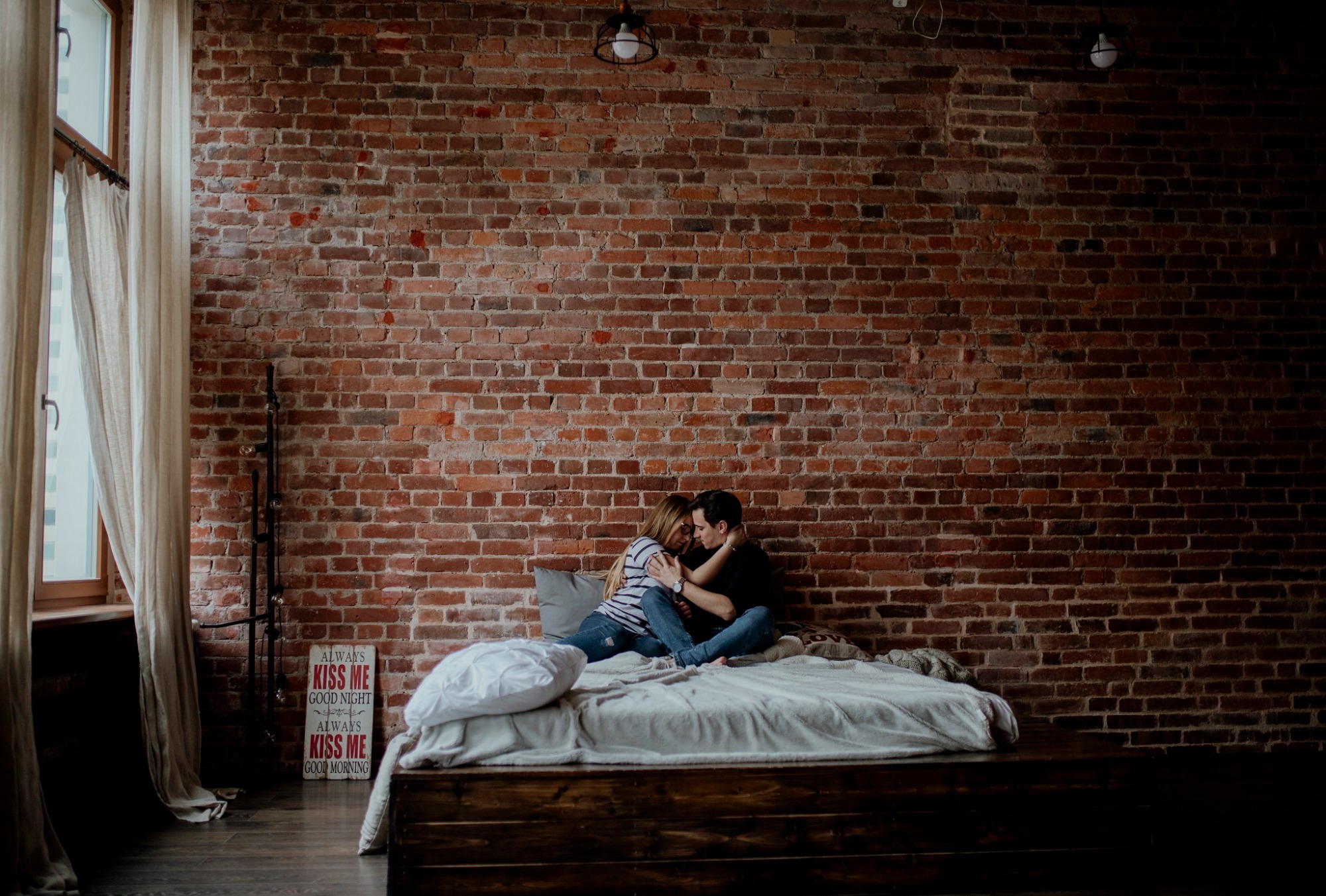 Para siedzi na łóżku na tle ceglanej ściany w loftowym wnętrzu, z widocznym oknem z zasłonami i dekoracyjnym napisem 'Kiss me good night'.