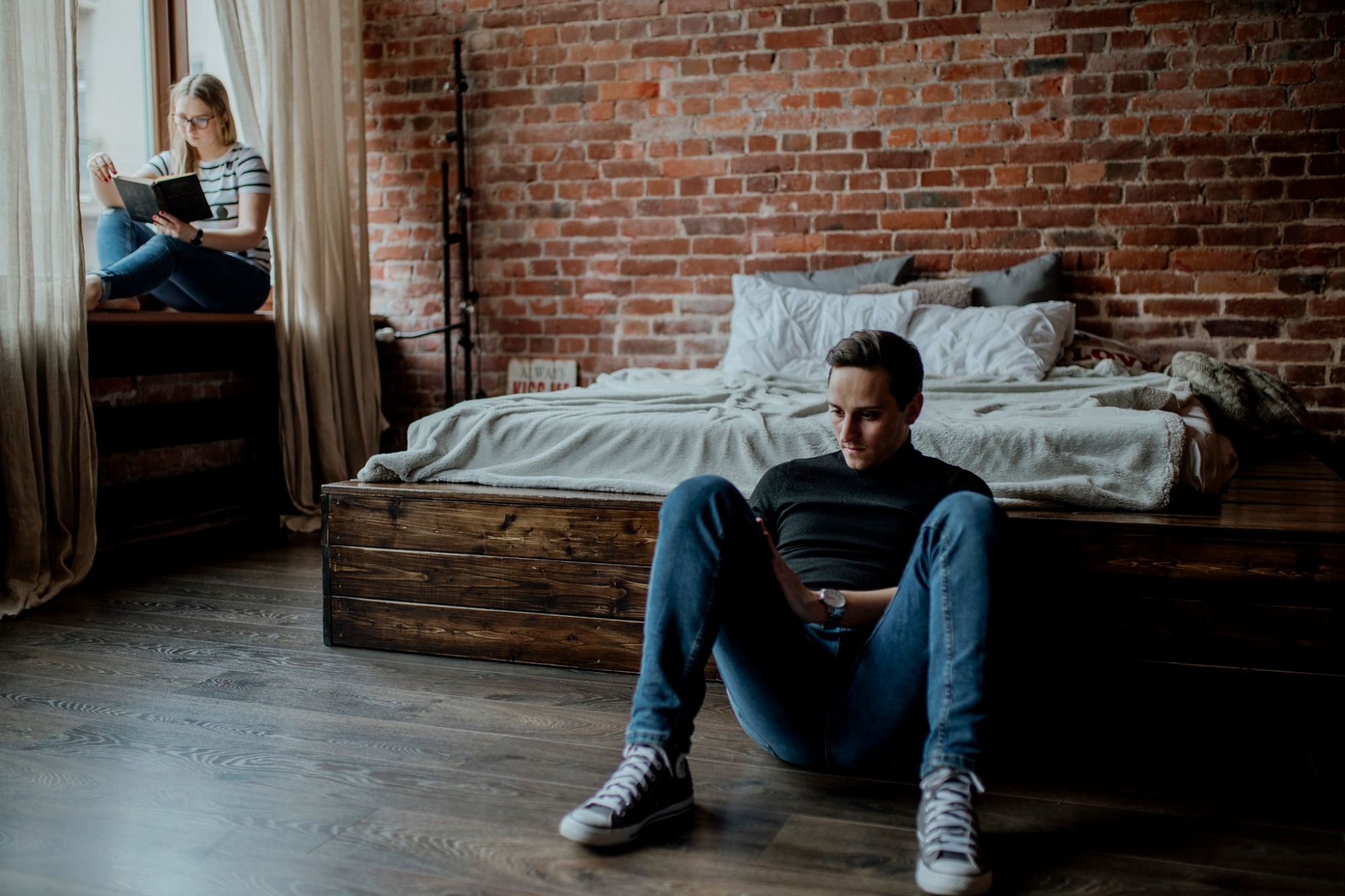 Młody mężczyzna siedzi na podłodze w loftowym wnętrzu z ceglaną ścianą, spoglądając na zegarek; w tle kobieta czyta książkę siedząc na parapecie okna.