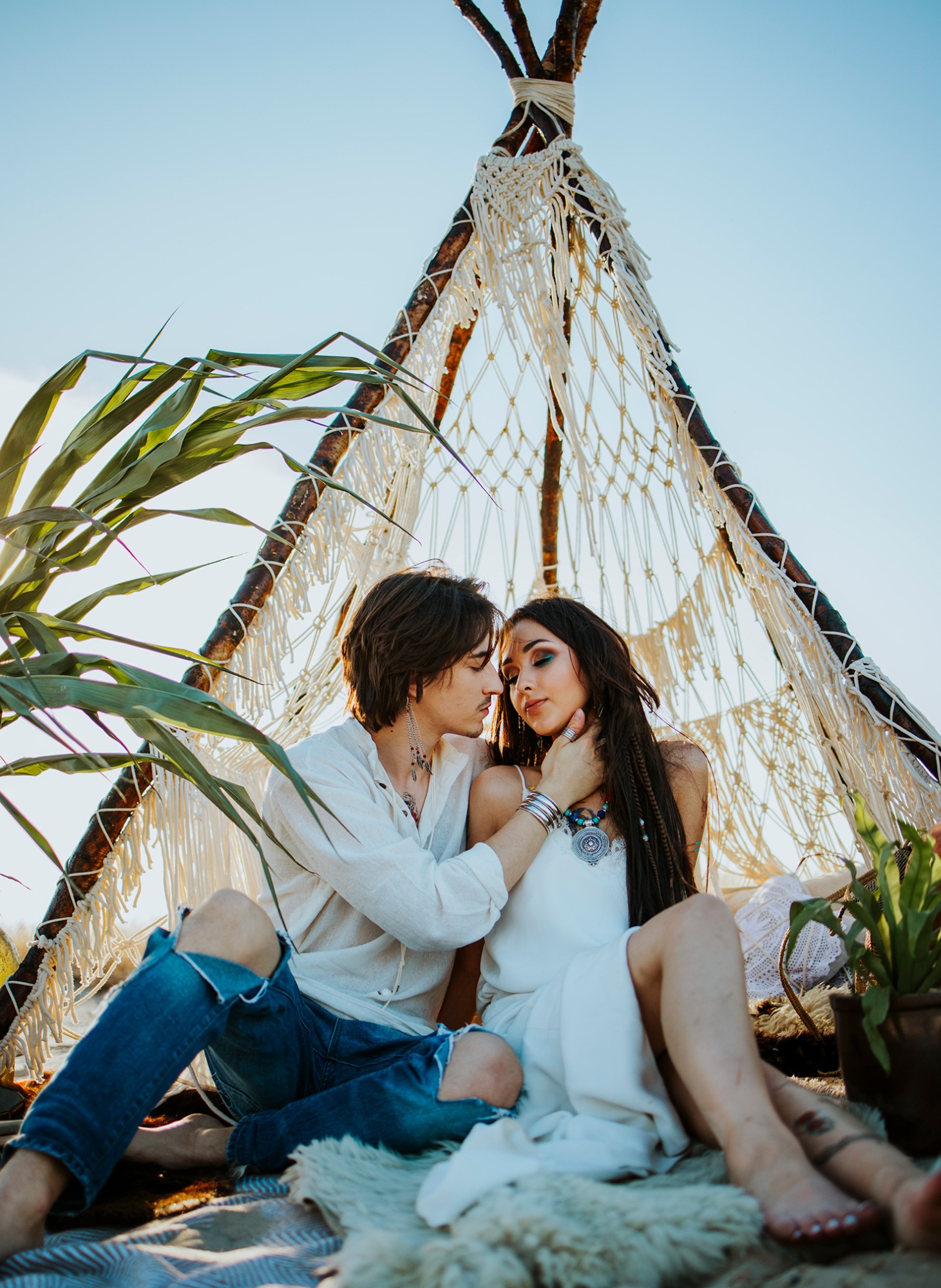 Para w stylu boho siedzi w tipi z makramy na tle nieba, dekoracje z roślin i poduszek, on w podartych jeansach, ona w białej sukience z biżuterią.