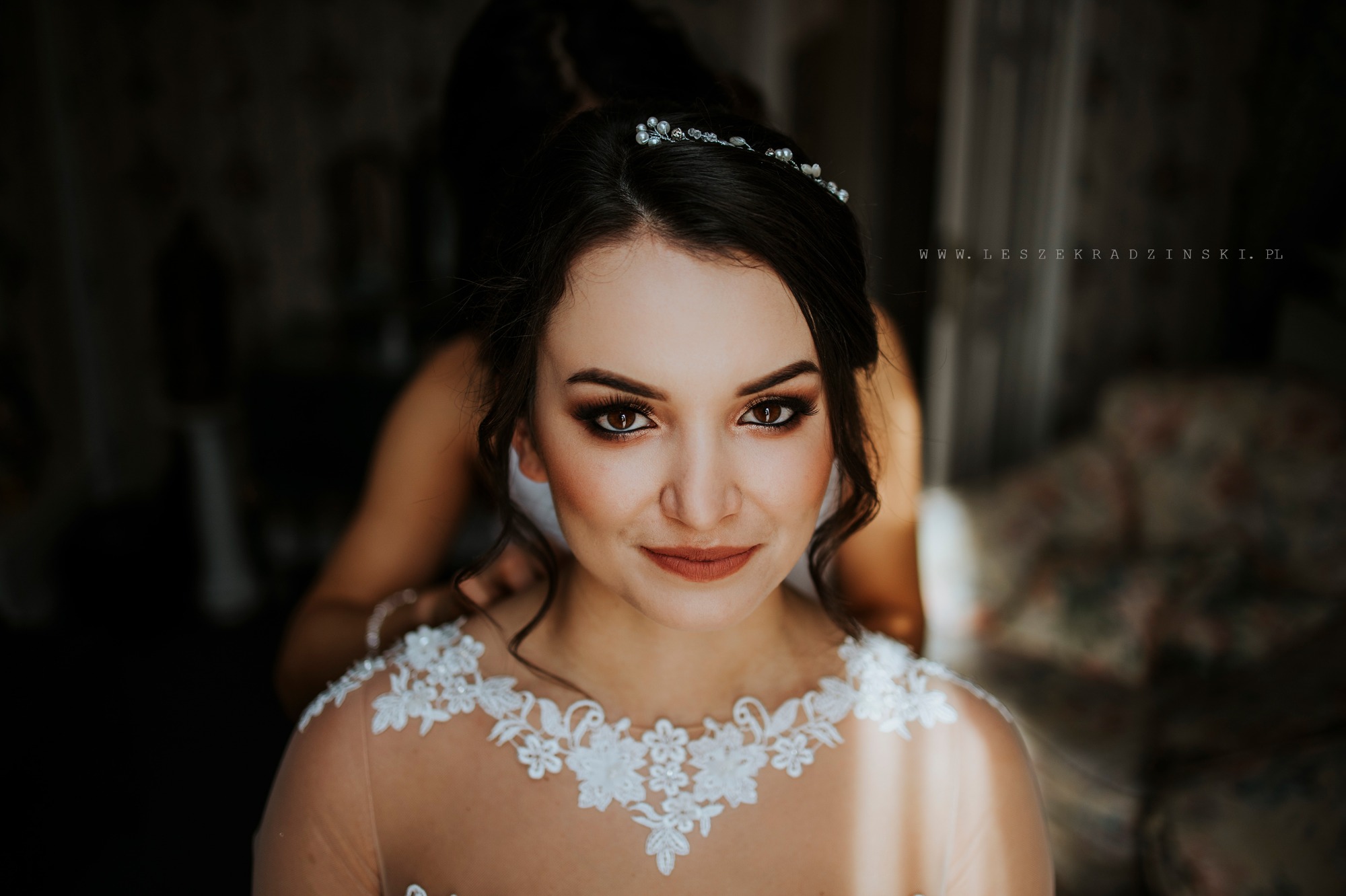 Portret panny młodej w delikatnej sukni z koronkowymi zdobieniami, z tiarą we włosach, podczas przygotowań do ślubu, z osobą pomagającą jej z tyłu.