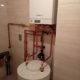 Biały piec gazowy Viessmann z miedzianą instalacją grzewczą i zasobnikiem ciepłej wody w łazience wyłożonej beżowymi płytkami.